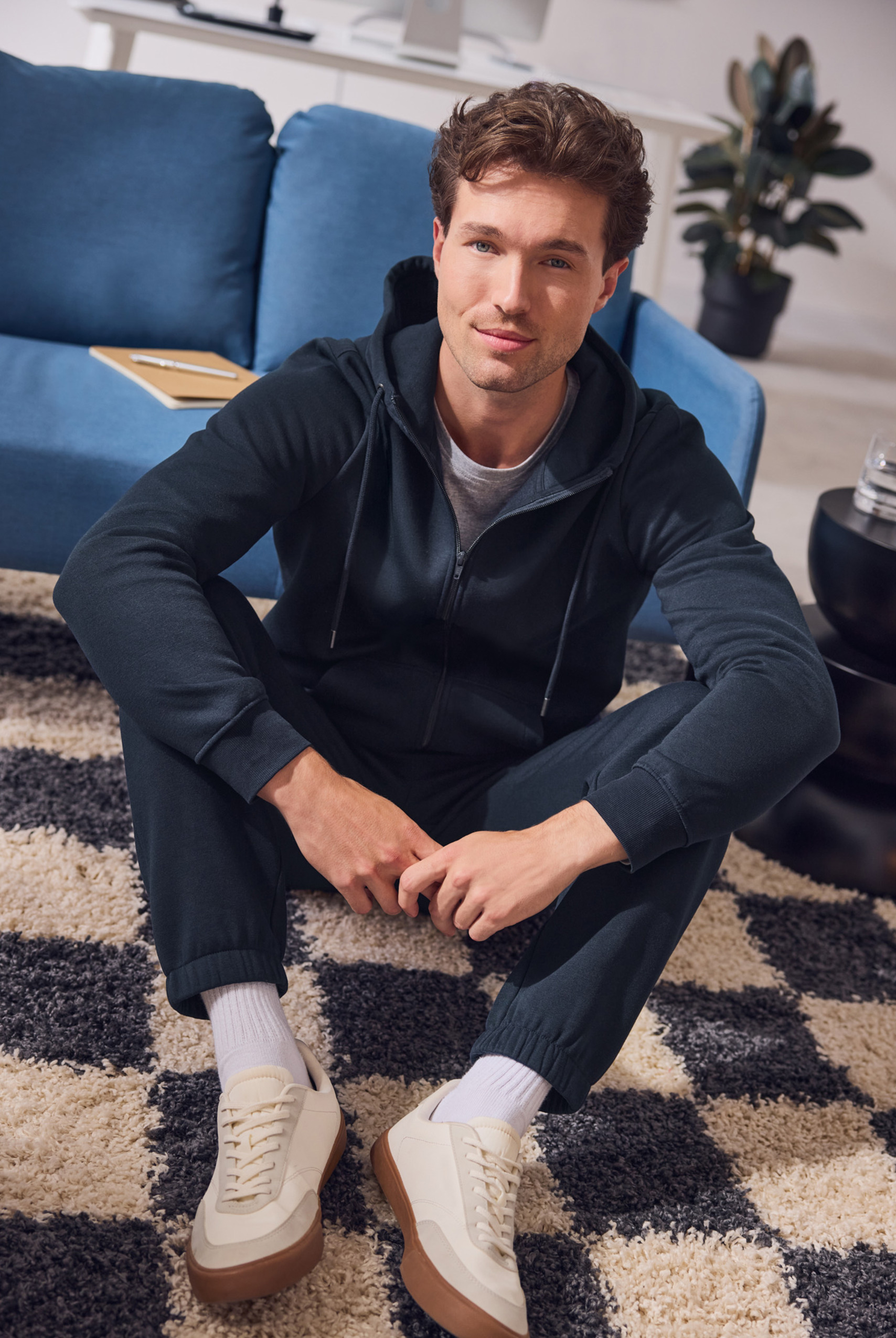 Ein Mann in einem dunkelblauen Kapuzenpullover und einer Jogginghose sitzt auf einem karierten Teppich.