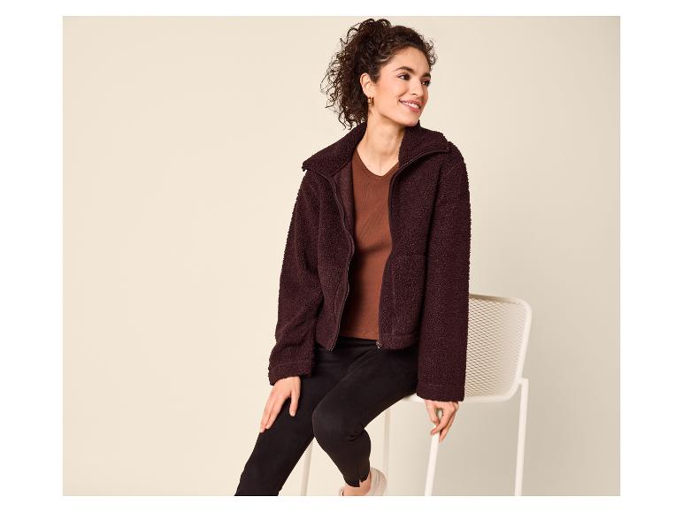 Frau in brauner Teddyfleecejacke, braunem Top und schwarzen Leggings, auf einem Stuhl sitzend.