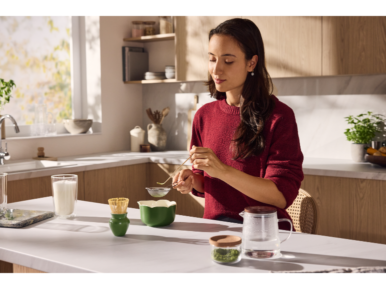Eine Frau bereitet Matcha-Tee in der Küche mit grünem Geschirr zu.