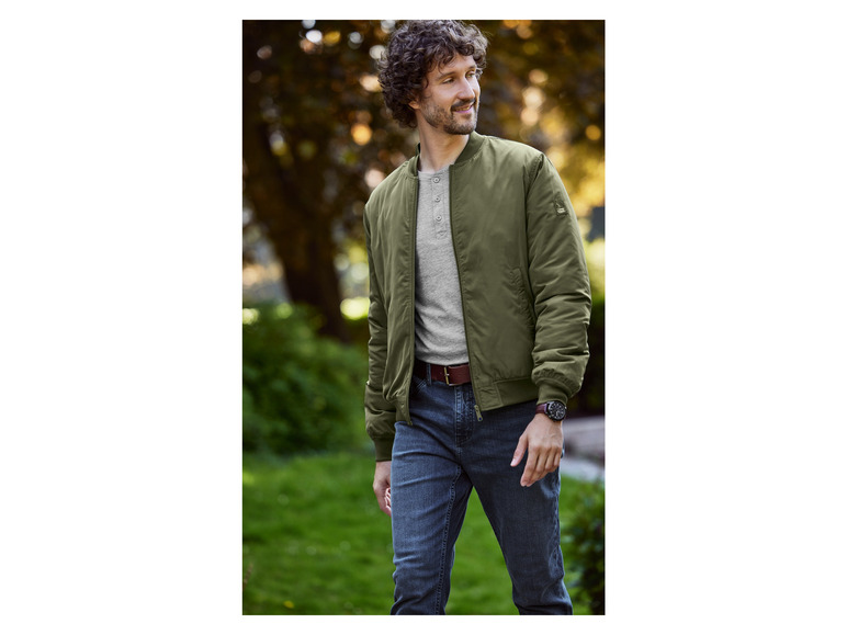 Mann trägt eine grüne Bomberjacke, Jeans und eine Uhr.
