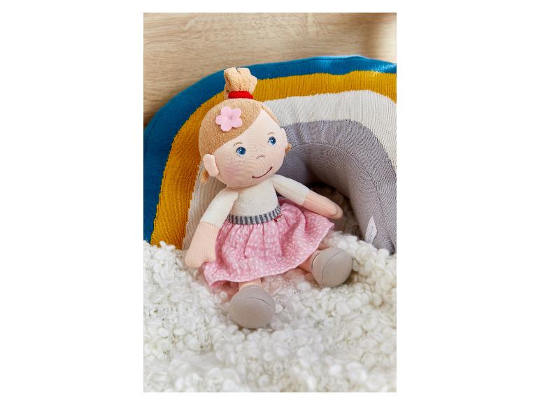 Eine Plüschpuppe mit rosa Rock und Regenbogenkissen auf einem weißen flauschigen Teppich.