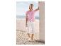 Frau in rosa perforiertem T-Shirt und weißen Caprihosen, an eine Wand gelehnt mit Meerblick.