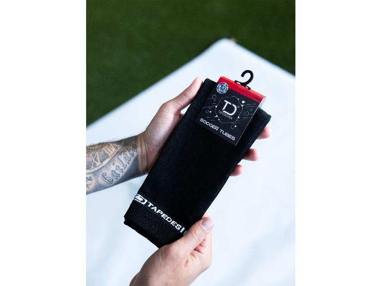 Hände halten schwarze Tapedesign Fußballstutzen mit dem Etikett „SOCCER TUBES“.