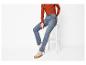 Frau in rostfarbenem Rippen-Top, blauen Jeans und beigen High-Top-Sneakern, sitzt auf einem weißen Hocker.