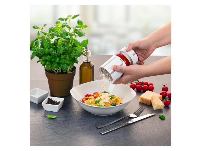 Eine Person reibt Käse über einen Teller Pasta mit Kirschtomaten und Basilikum, mit Olivenöl und Gewürzen.