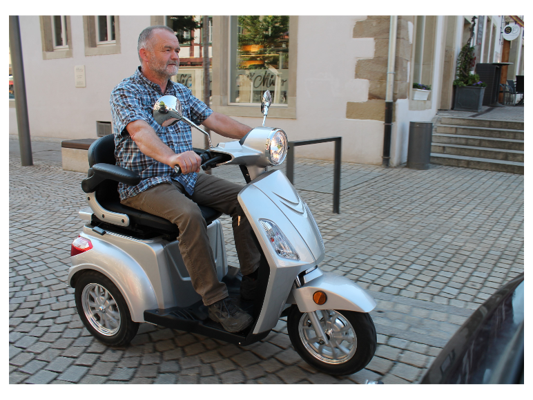 Ein Mann mittleren Alters fährt einen silbernen Dreiradroller auf einer Kopfsteinpflasterstraße.