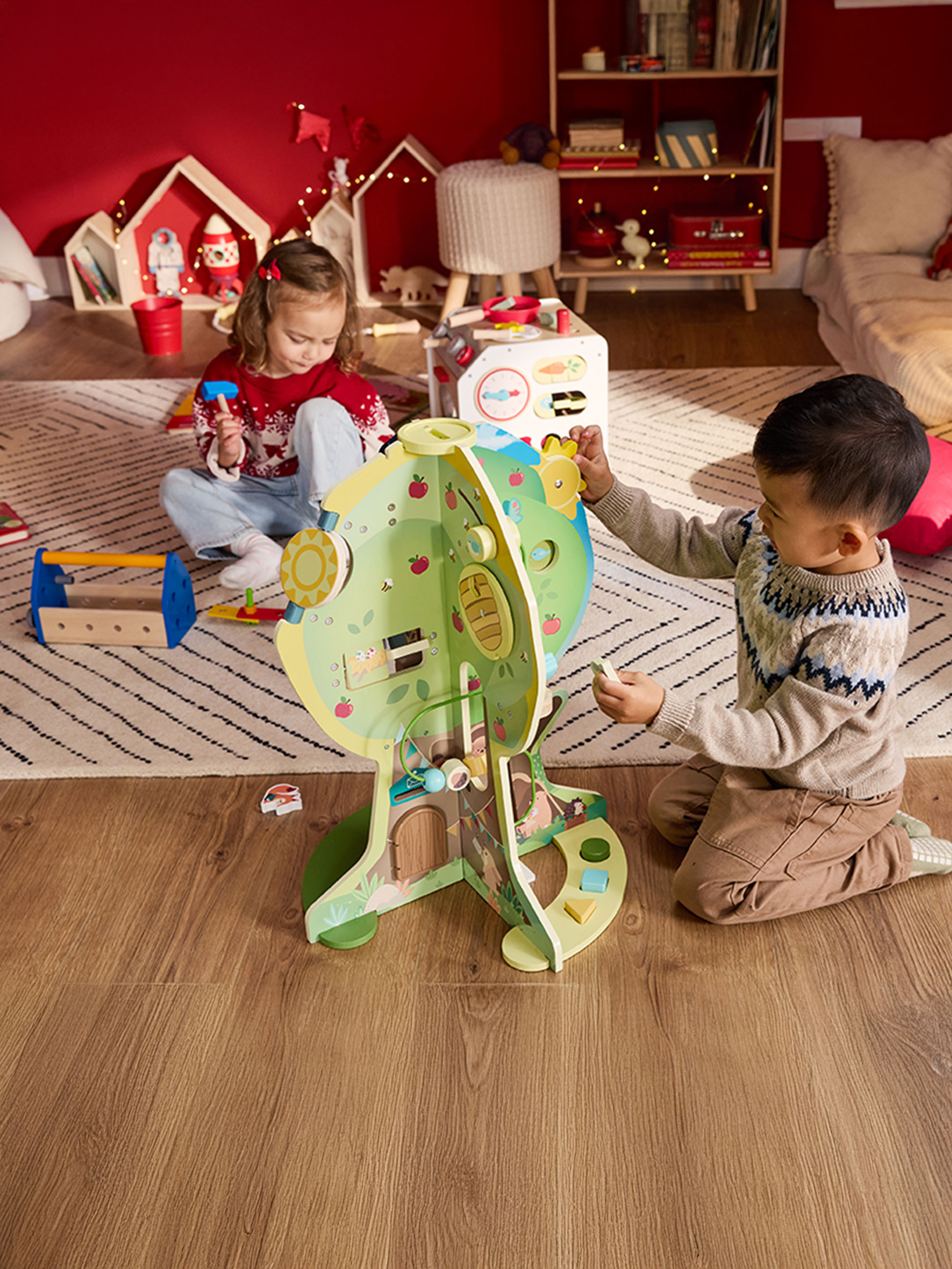 Zwei Kinder spielen mit einem Holzpuppenhaus und einem Werkzeugkasten in einem festlich dekorierten Zimmer.