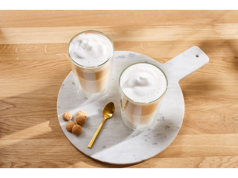 Zwei Gläser geschichteter Latte Macchiato mit Schaum, ein goldener Löffel und Kekse auf einem Marmorbrett.