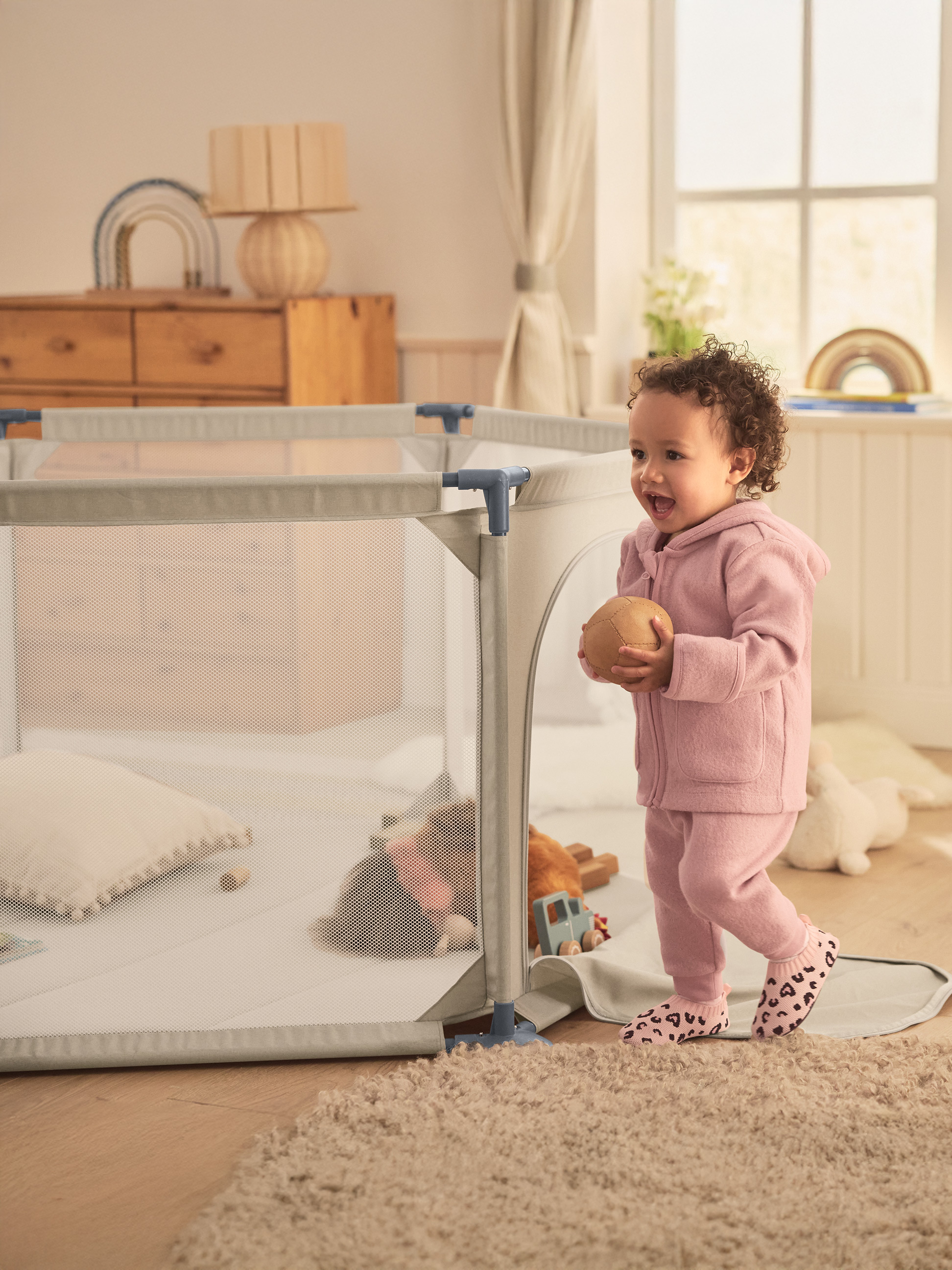 Kleinkind in rosa Kleidung hält Ball, steht neben Laufstall mit Spielzeug.
