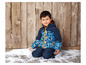 Ein Junge in einer blauen Skijacke mit geometrischem Muster sitzt im Schnee.