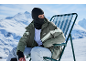 Mann mit Sturmhaube und grüner Skijacke sitzt auf einem Stuhl in verschneiten Bergen.
