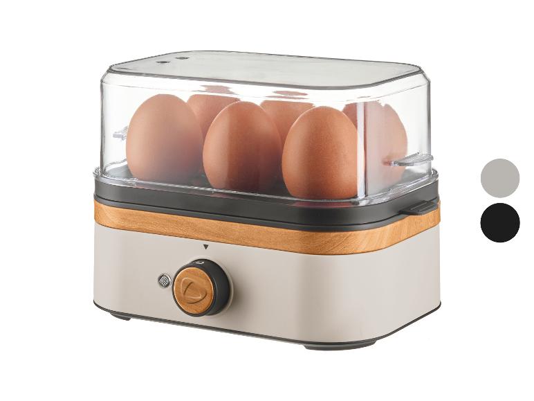 Eierkocher mit transparentem Deckel, braunen Eiern, hellem Holz und grauem Sockel.