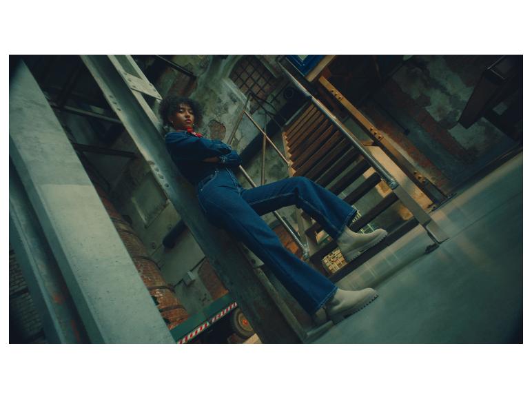 Eine Frau in Jeans-Overall und hellen Stiefeln posiert auf Industriestufen.