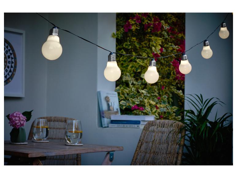 Lichterkette beleuchtet eine gemütliche Terrasse mit Holztisch und Korbstühlen.