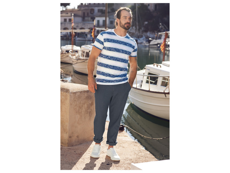 Mann in einem gestreiften T-Shirt, blauen Hosen und weißen Sneakers in einem Hafen.