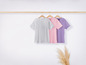 Drei T-Shirts in Grau, Rosa und Lila hängen an einem Holzbügel.