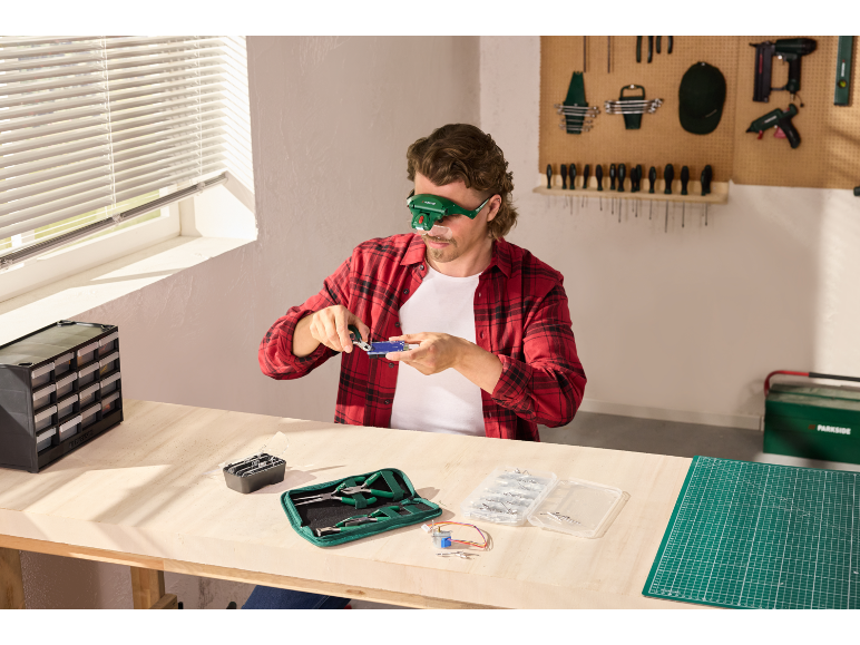Mann mit Lupenbrille arbeitet an einer Elektronikplatine, umgeben von Werkzeugen und Komponenten.