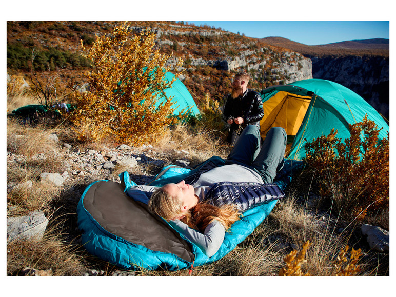 Eine Frau schläft in einem blauen Schlafsack neben einem Zelt in den Bergen.