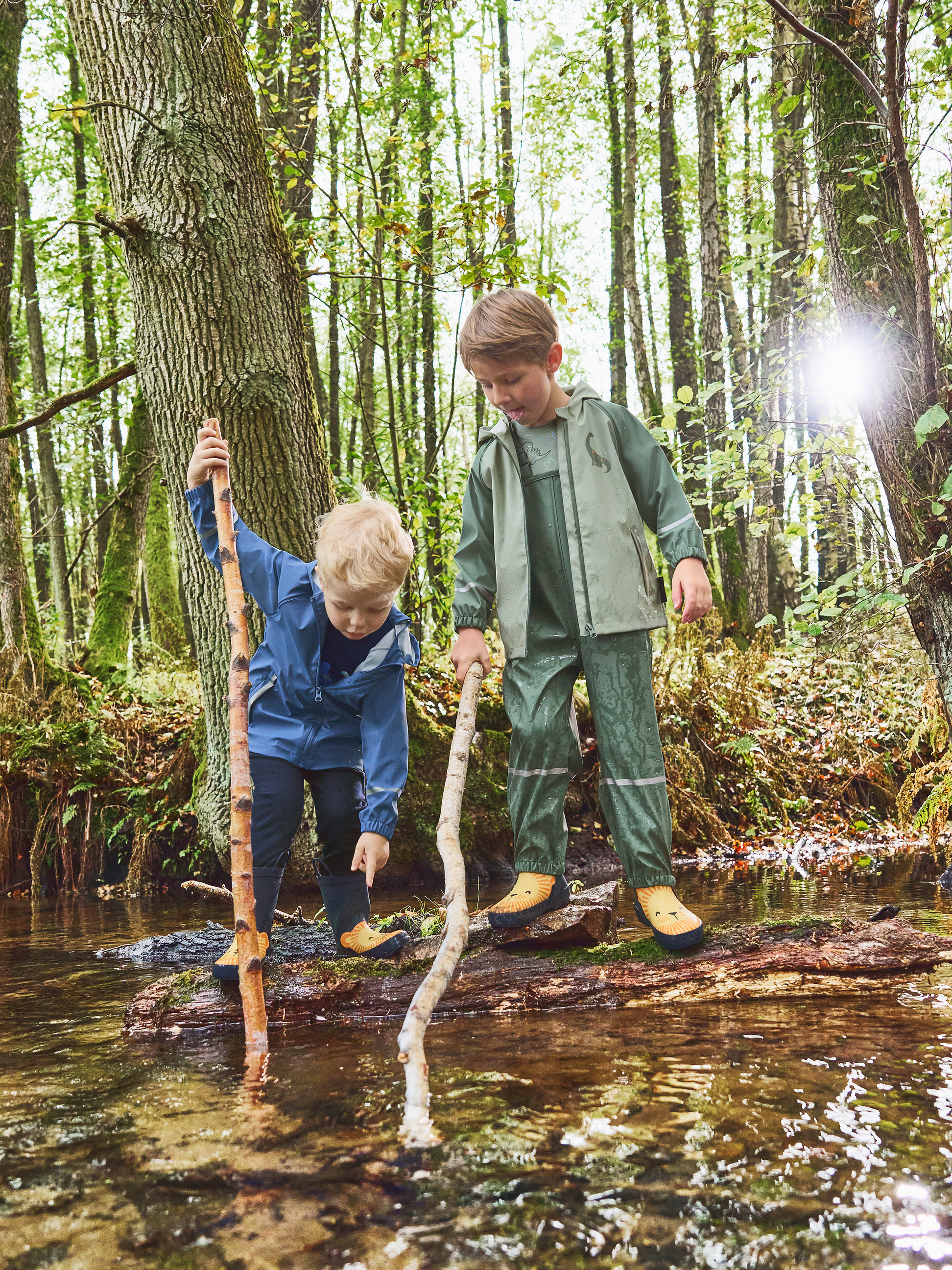 Zwei Kinder in Regenmänteln und Gummistiefeln spielen mit Stöcken in einem Wald Bach.
