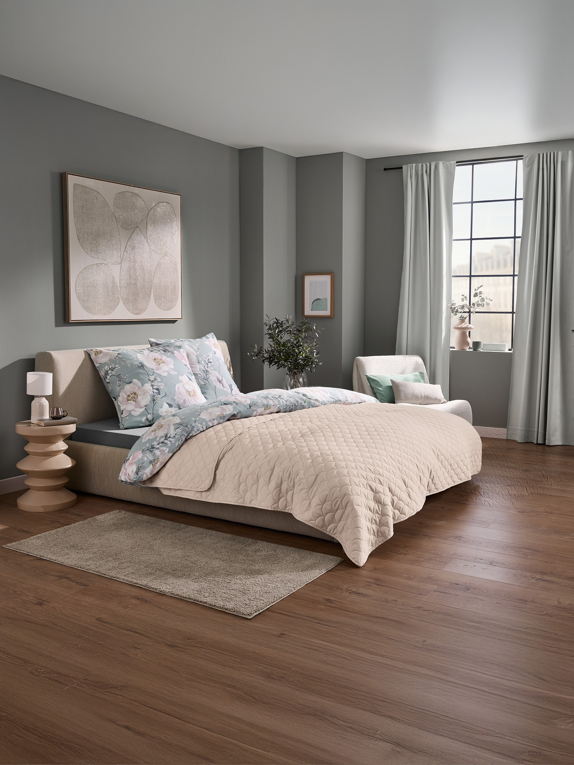 Schlafzimmer mit Bett, floraler Bettwäsche und beiger Steppdecke, sowie einem Fenster mit Vorhängen.