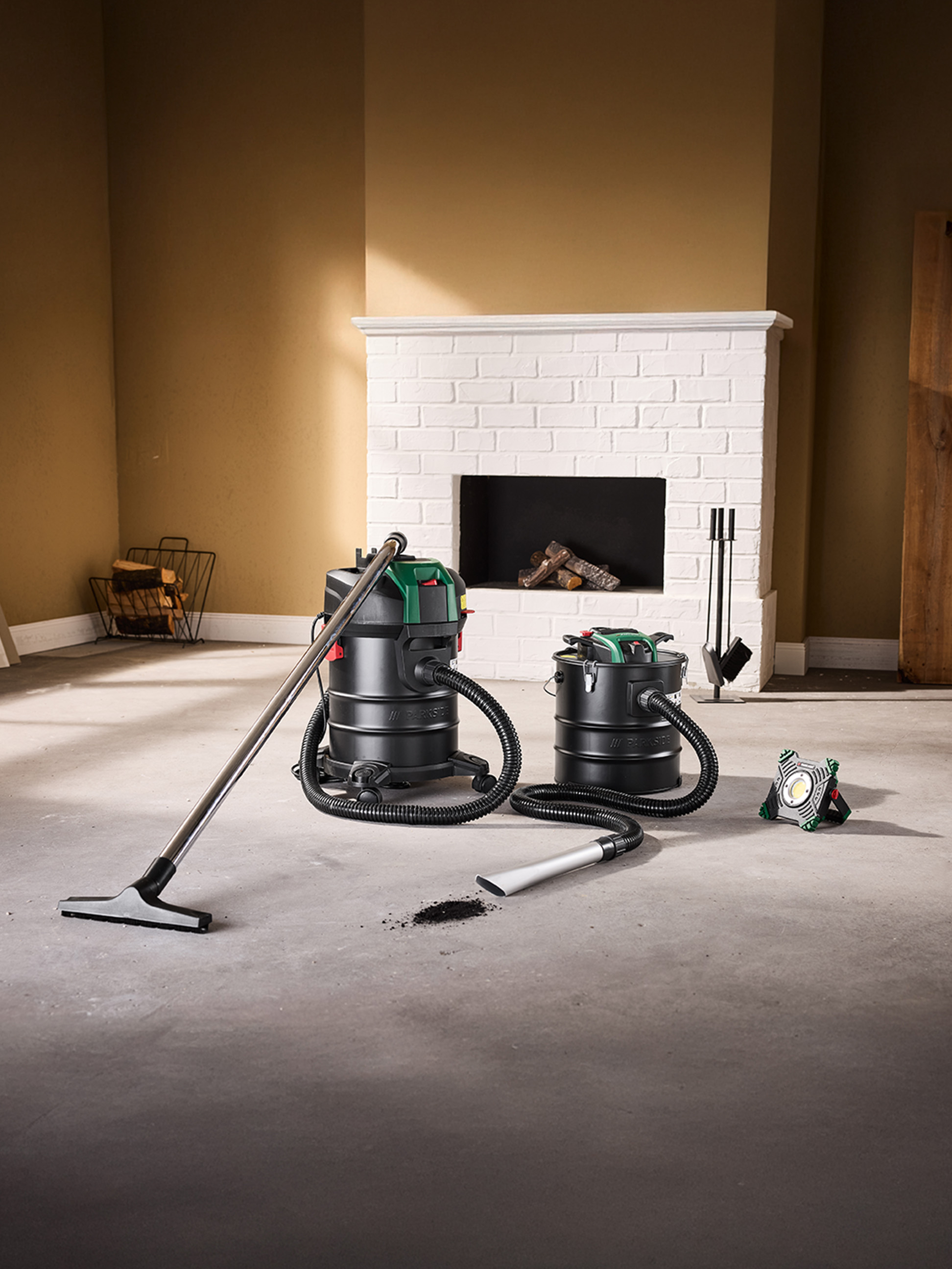 Zwei Aschesauger und eine Arbeitsleuchte auf Betonboden vor einem Kamin.
