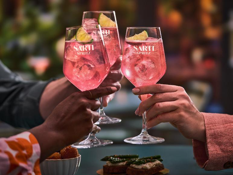 Drei Personen stoßen mit rosa Spritz-Cocktails an, garniert mit Limette, mit Snacks auf dem Tisch.