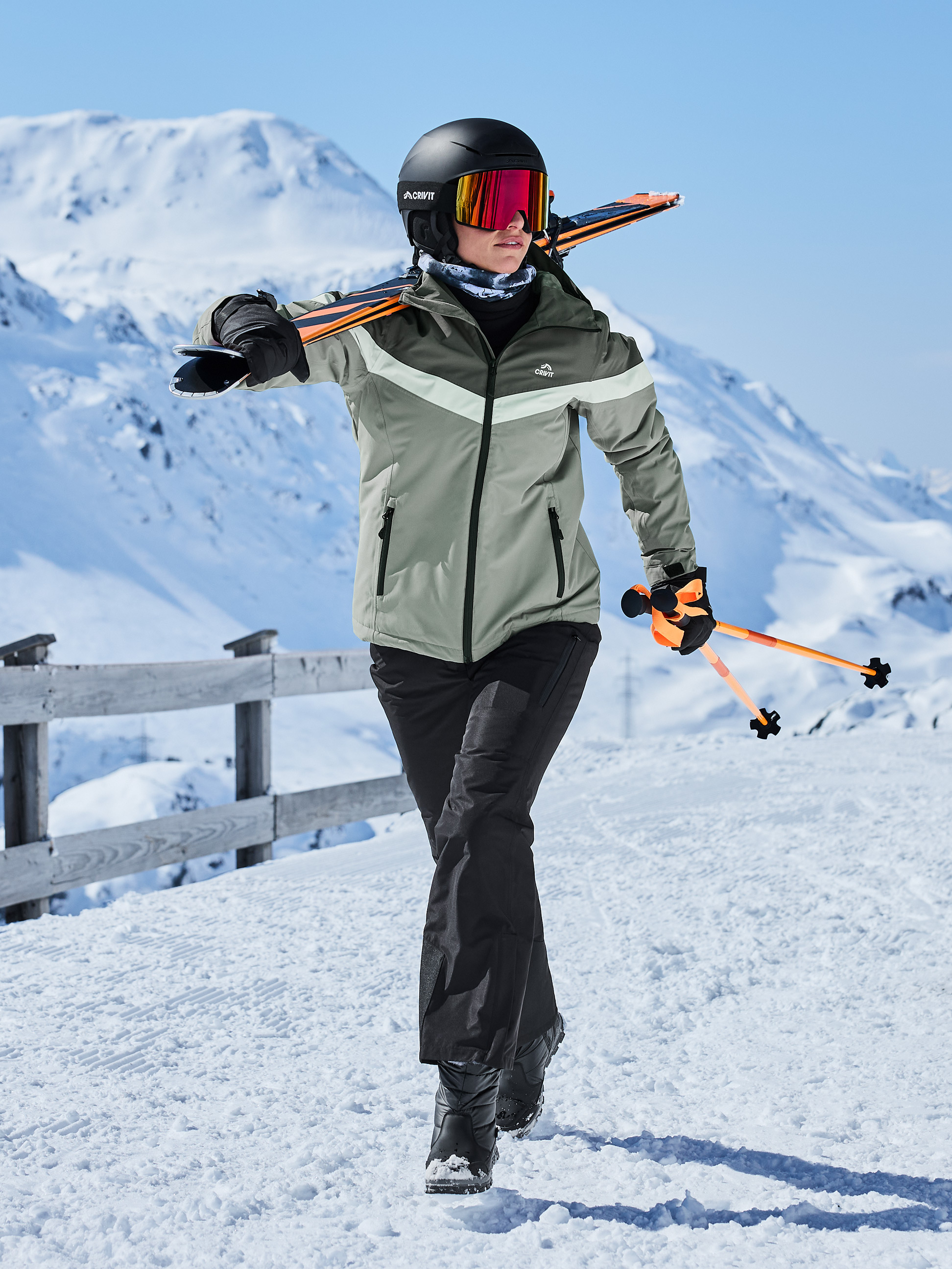 Frau in Skijacke, Hose, Helm, Brille und Skiern über der Schulter im Schnee.