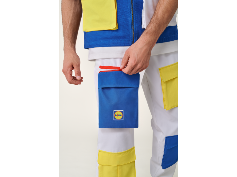 Detail eines Mannes in weißer Weste und Hose mit blauen und gelben Taschen, der eine Tasche mit rotem Reißverschluss öffnet.