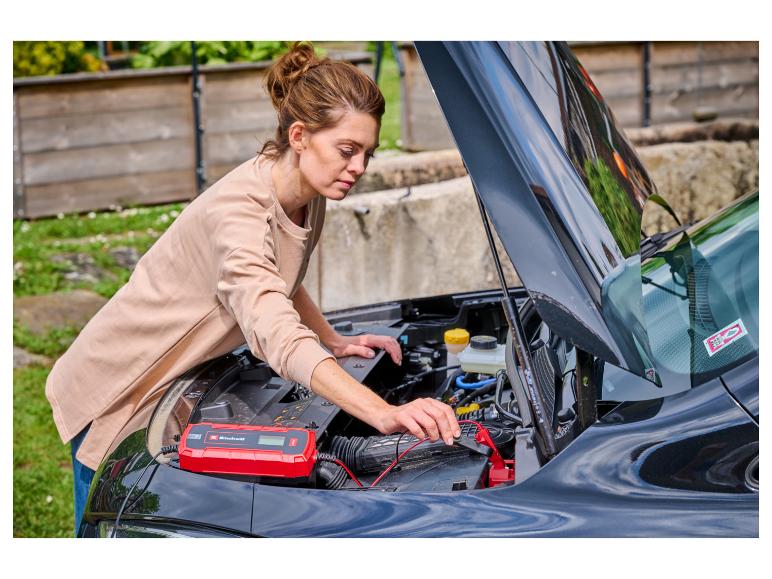 Frau lädt Autobatterie mit rotem Ladegerät unter geöffneter Motorhaube.