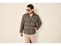 Mann in dunkelgrauer Pufferjacke, hellbeigem Pullover und hellbeiger Hose.