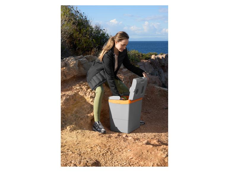 Frau öffnet eine grau-orange elektrische Kühlbox auf felsigem Gelände am Meer.