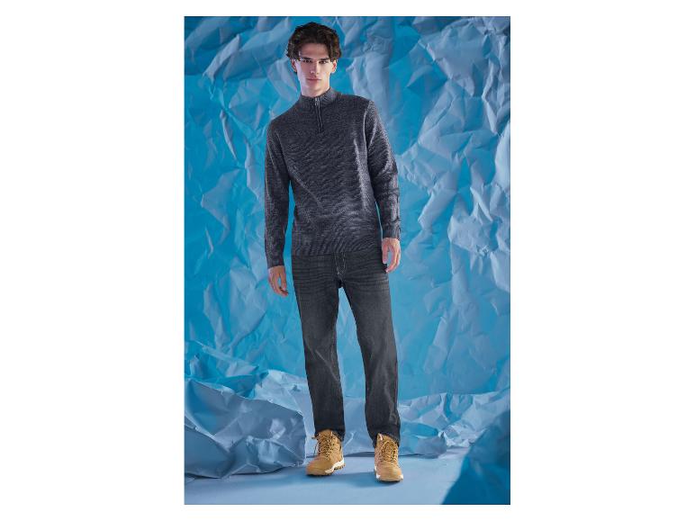 Ein Mann in grauem Pullover und dunklen Jeans vor blauem Hintergrund.