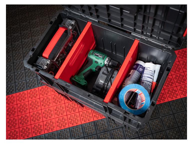 Offene Werkzeugkiste mit Bohrmaschine, Ladegerät, Dichtmitteln und Klebeband auf gemustertem Boden.