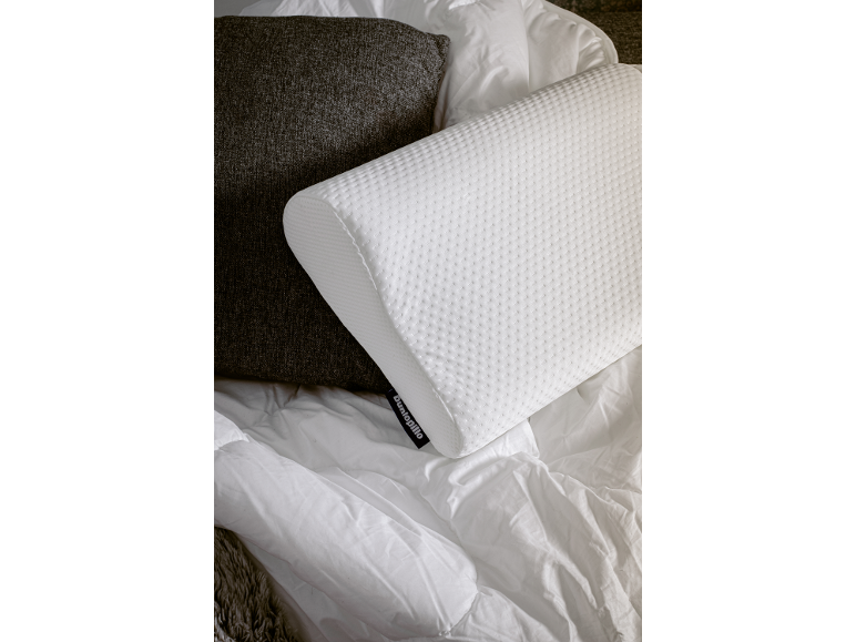 Weißes ergonomisches Kissen mit strukturierter Hülle auf einem Bett mit dunkelgrauem Kissen und weißer Bettdecke.