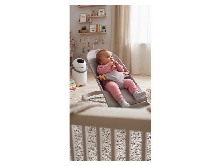 Baby in einer grauen Stoffwippe in einem Kinderzimmer mit Holzregalen und Dekoration.
