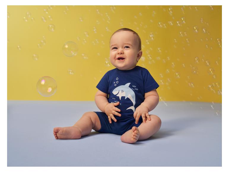 Lächelndes Baby im blauen Strampler mit Hai, umgeben von Seifenblasen.