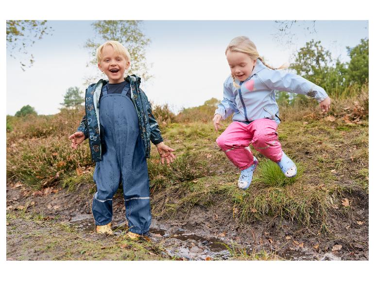 Zwei glückliche Kinder in wasserdichten Jacken und Latzhosen spielen in schlammigen Pfützen.