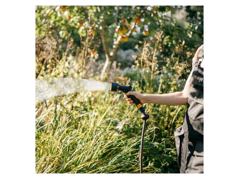 Person bewässert Pflanzen mit Gartenschlauch und Spritzpistole in einem sonnigen Garten.
