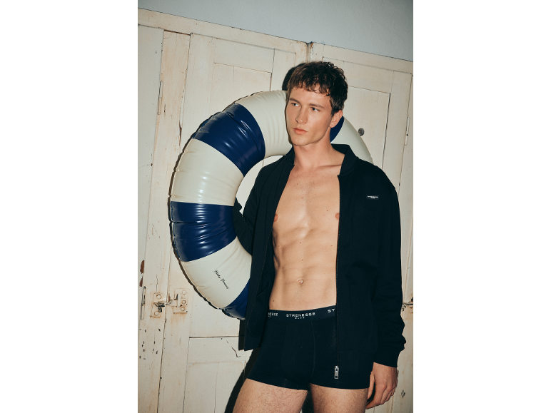 Mann in schwarzen Boxershorts und offener Jacke hält einen Schwimmreifen.