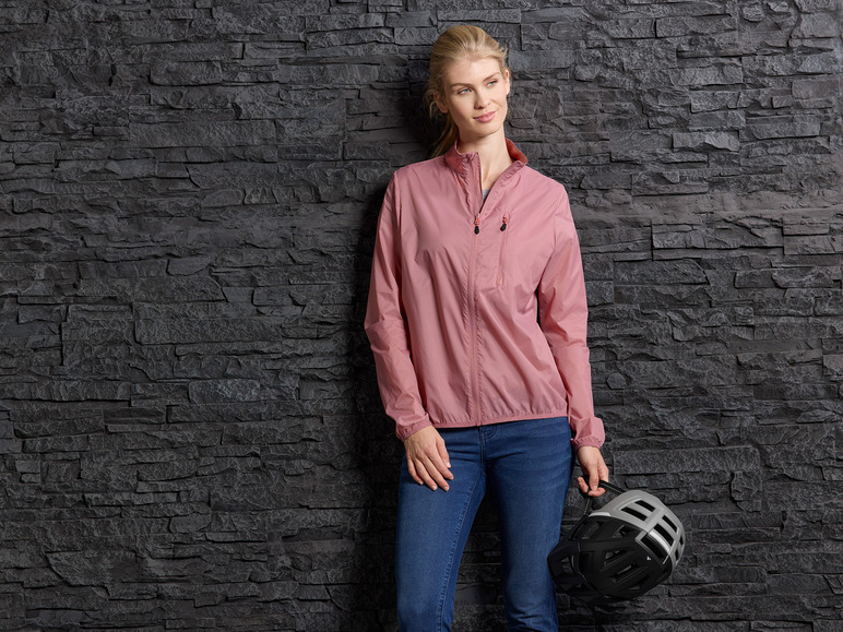 Eine Frau in einer rosafarbenen Windjacke und blauen Jeans hält einen Fahrradhelm in der Hand.