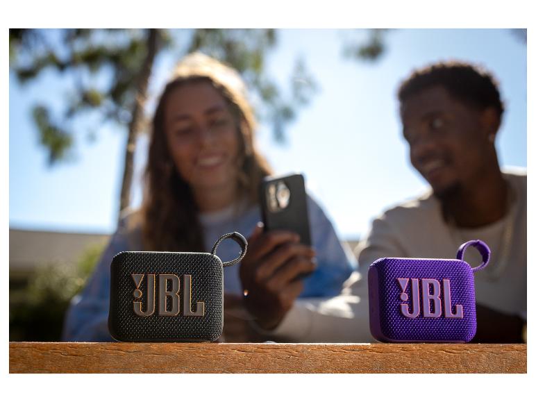 Zwei JBL-Lautsprecher auf einem Holztisch, im Hintergrund lächelnde Menschen.