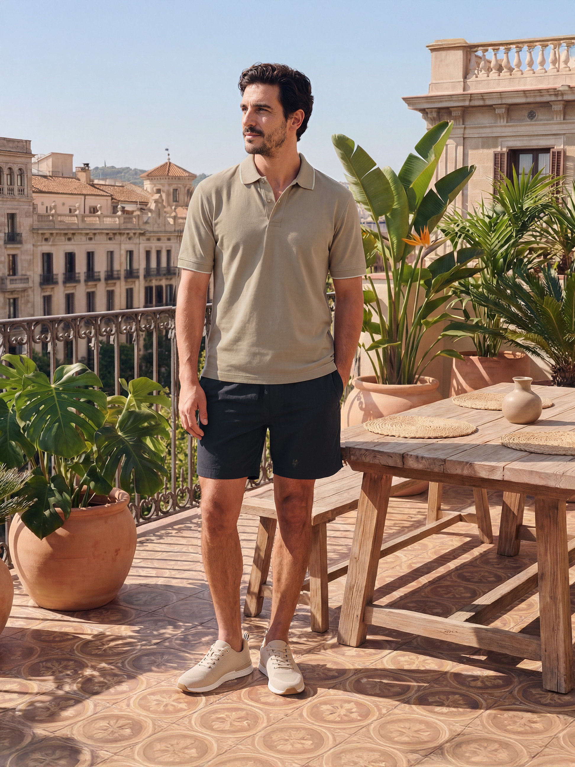 Mann in beigem Poloshirt und schwarzen Shorts auf sonniger Terrasse mit Stadtblick.