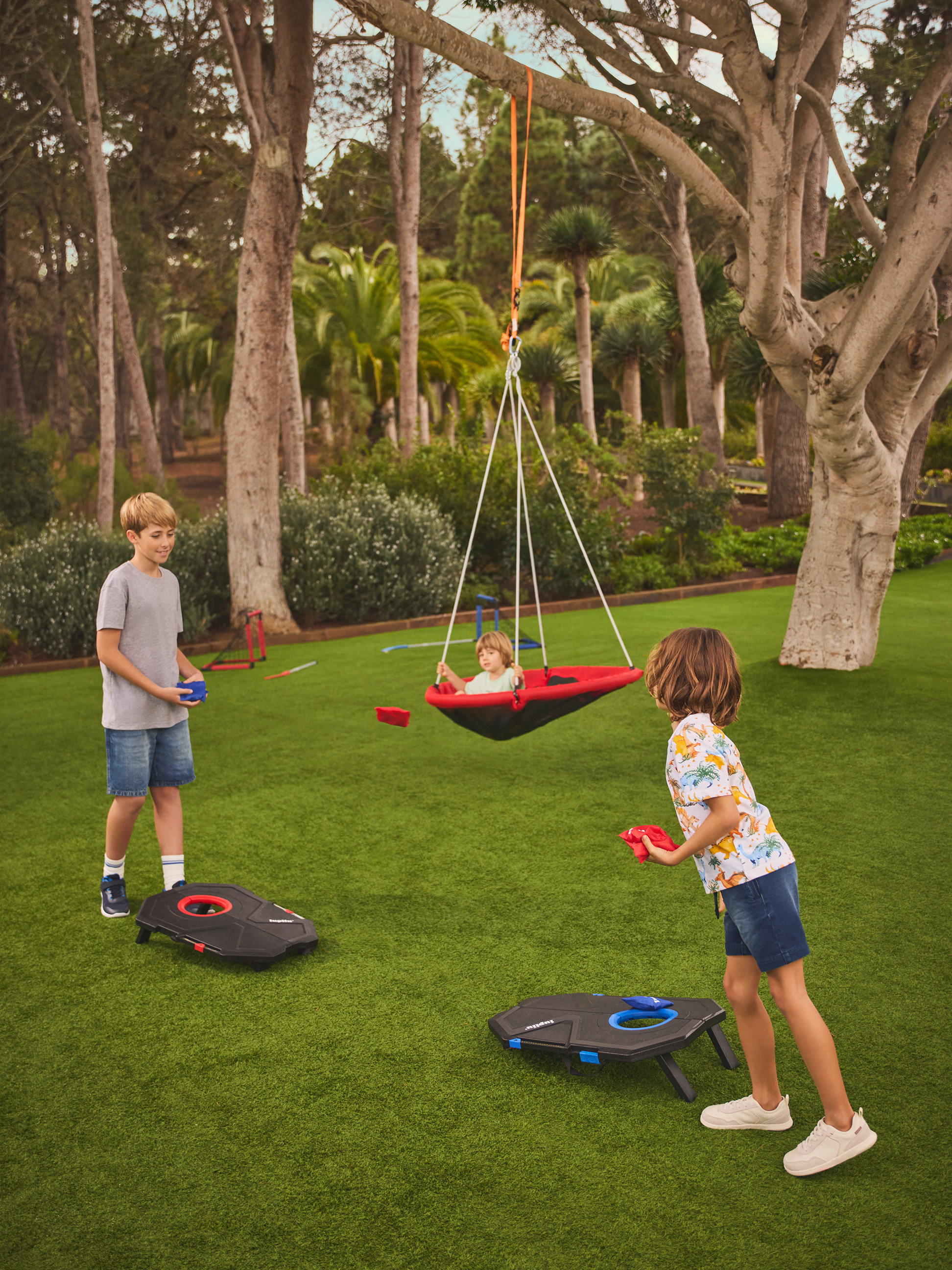 Zwei Kinder spielen Cornhole auf dem Rasen, während ein drittes in einer Nestschaukel schaukelt.