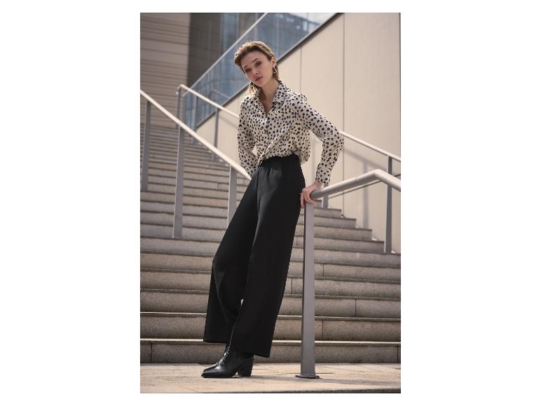 Frau in Leopardenmuster-Hemd und schwarzer Hose, an einem Geländer lehnend.
