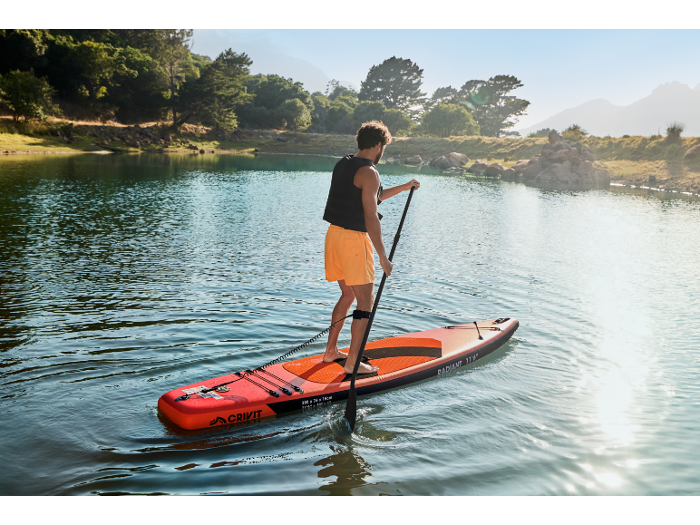 Mann paddelt auf einem Stand-Up-Paddle-Board auf einem See, umgeben von Bäumen und Bergen.