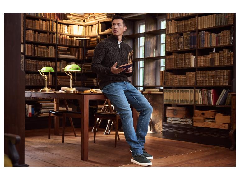 Mann in Strickpullover und Jeans sitzt an einem Bibliothekstisch und liest ein Buch.