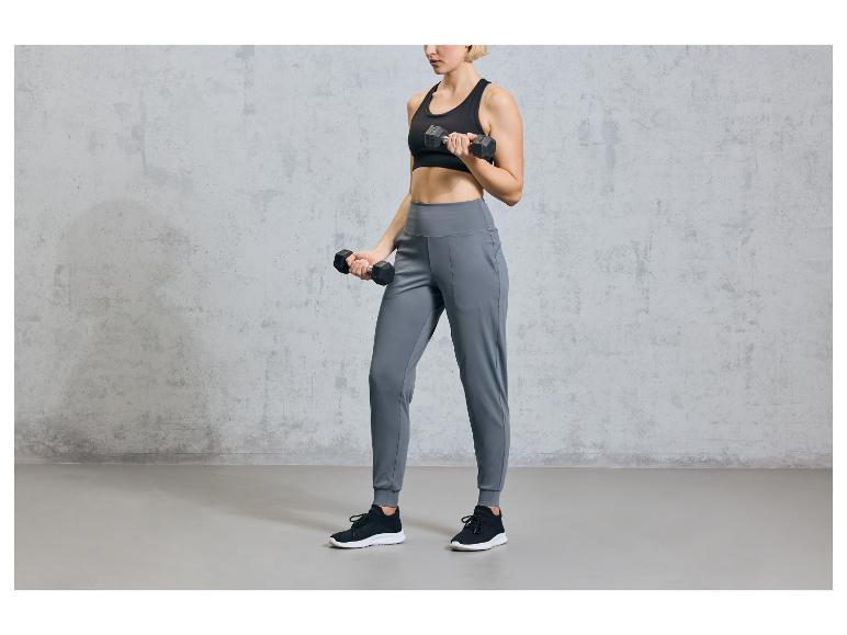 Eine Frau trägt schwarze Sportkleidung und graue Jogginghosen, während sie Kurzhanteln benutzt.