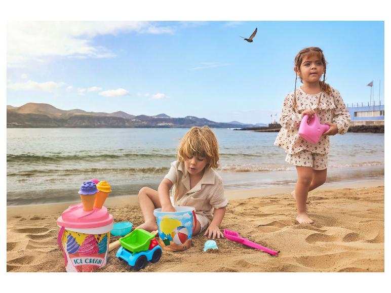 Zwei Kinder spielen mit Sandspielzeug am Strand, mit Meer und Bergen im Hintergrund.