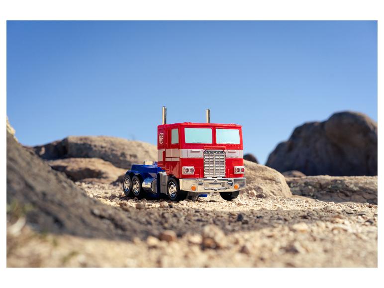 Ein roter und blauer Optimus Prime Spielzeug-LKW auf felsigem, sandigem Gelände unter klarem Himmel.