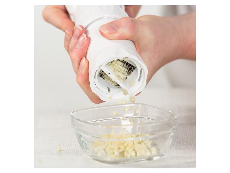 Hände verwenden einen weißen Knoblauchhacker, um Knoblauch in eine Glasschale zu zerkleinern.